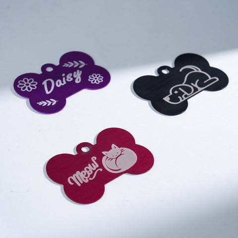 Three Creality Falcon Large Multicolor Anodized Aluminum Dog Tags are shown: a purple floral "Daisy" bone tag, a black etched bone tag, and a red tag engraved with "Meow" and a cat image, all displayed on a white surface.
